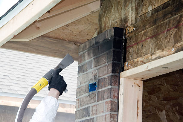 A construction worker uses a power tool to work on a brick chimney during what appears to be a home renovation or construction project with exposed wooden framing visible.