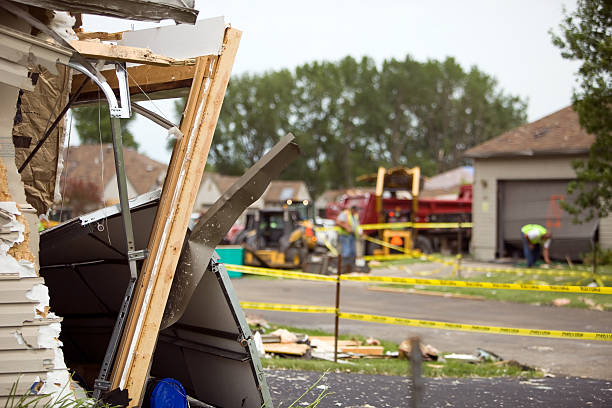 A severely damaged house with collapsed structural beams is cordoned off with yellow caution tape while emergency vehicles and workers respond to what appears to be a disaster scene in a residential neighborhood.