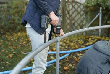 A person in dark clothing uses a power tool to work on metal railings or fencing in what appears to be an outdoor construction or maintenance project.