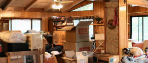 A wooden-paneled room with exposed beams is filled with stacks of moving boxes and covered furniture, indicating someone is in the process of packing up or moving out.